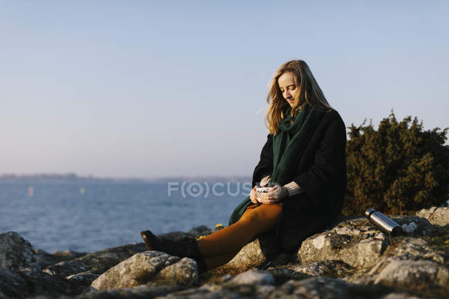 Woman holding mug sitting on rocks by sea — Stock Photo