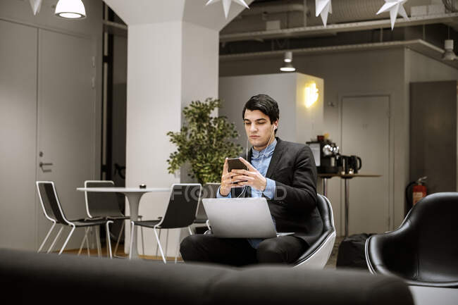 Молодий чоловік в навушниках використовує смартфон і ноутбук в офісі — стокове фото
