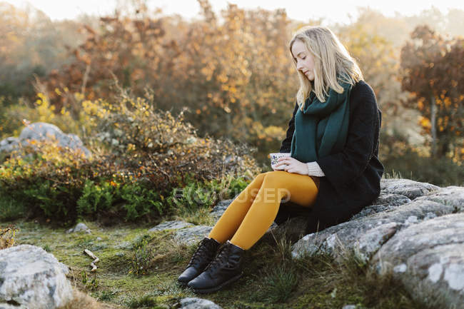 Woman holding mug sitting on rocks by bushes — Stock Photo
