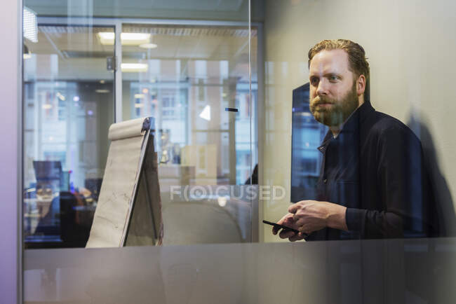 Un homme tenant un téléphone intelligent derrière une fenêtre de bureau — Photo de stock