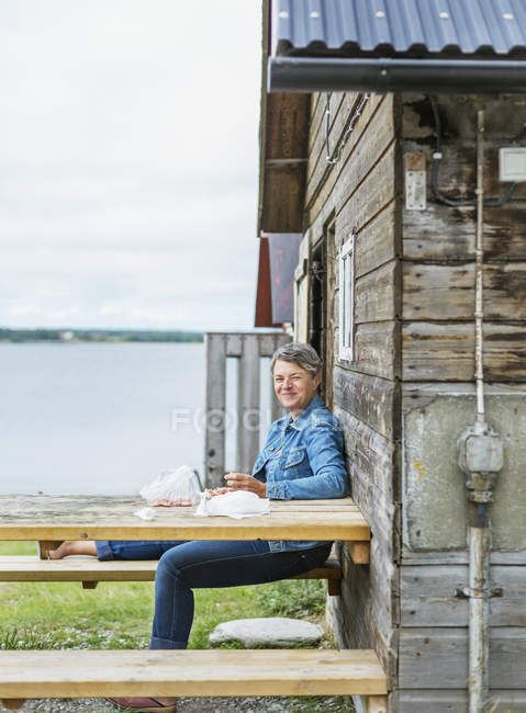Зрелая женщина сидит за столом для пикника рядом с бревенчатой хижиной — стоковое фото