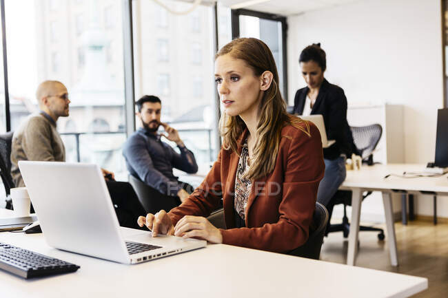 Empresária usando laptop na mesa enquanto trabalhava com colegas no escritório — Fotografia de Stock