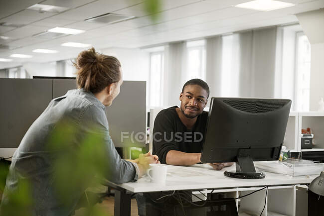 Smiling male coworkers sitting at desk and talking in office - foto de stock