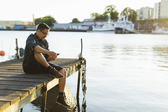 Mann sitzt auf Steg und benutzt Smartphone — Stockfoto