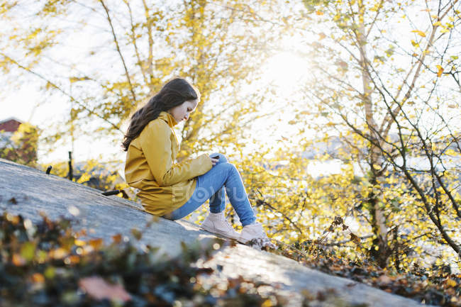 Ragazza che indossa impermeabile giallo utilizzando smart phone dagli alberi — Foto stock