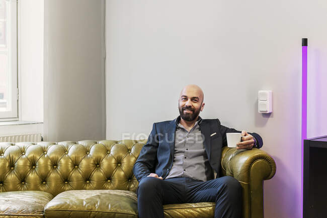 Homem segurando caneca enquanto sentado no sofá e sorrindo para a câmera — Fotografia de Stock