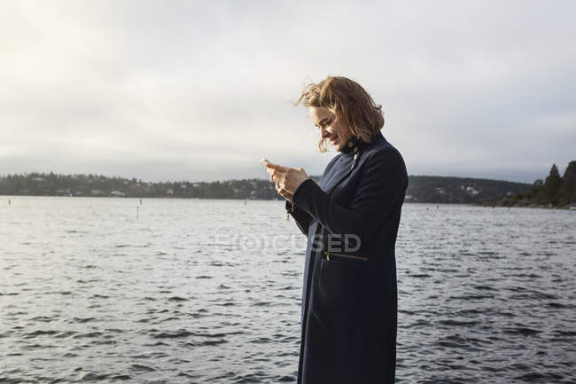 Young woman standing next to lake and holding cell phone — Stock Photo