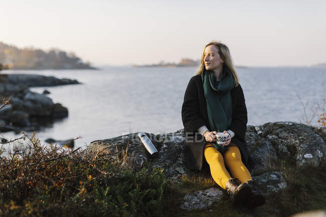Жінка тримає кухоль, сидячи на каменях біля моря — стокове фото