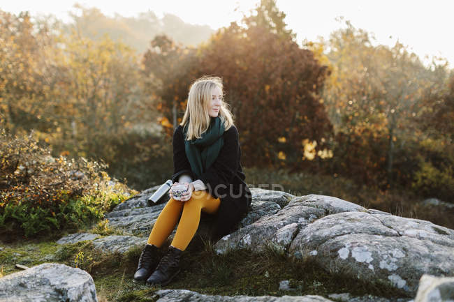 Mulher segurando caneca sentado em rochas por arbustos — Fotografia de Stock
