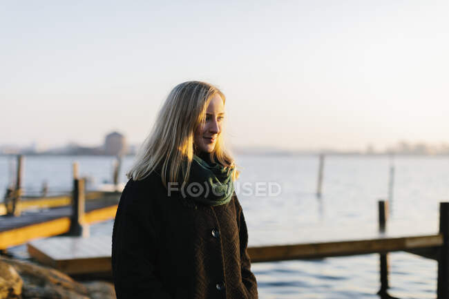 Молода жінка в пальто і шарфі біля моря на заході сонця — стокове фото