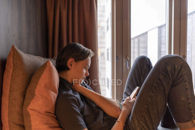 Adolescente menino inclinado em almofadas usando telefone inteligente — Fotografia de Stock
