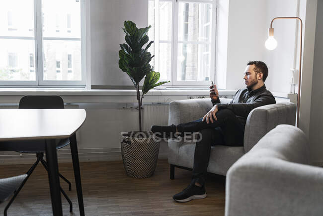 Uomo seduto in poltrona e con smartphone da lampada — Foto stock