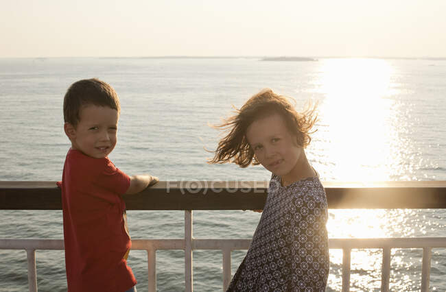 Hermano y hermana mirando a la cámara mientras se encuentra junto al mar al atardecer. - foto de stock