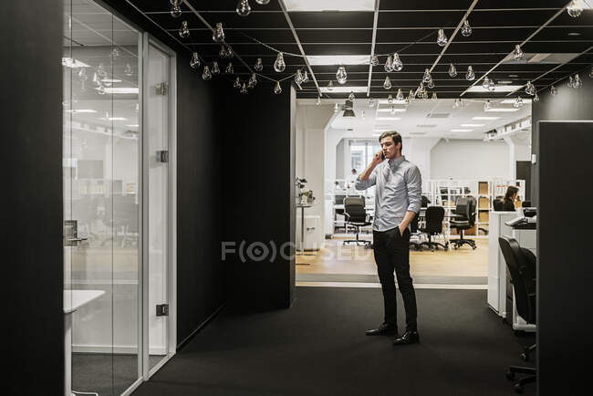 Молодой человек стоит с рукой в кармане и разговаривает по телефону в офисе — стоковое фото