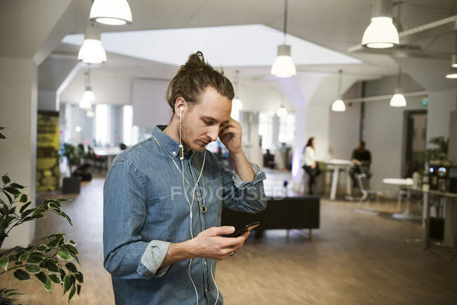 Focalizzato giovane uomo con gli auricolari con smartphone in ufficio — Foto stock