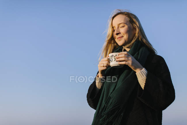 Mujer joven con los ojos cerrados sosteniendo taza contra el cielo despejado - foto de stock