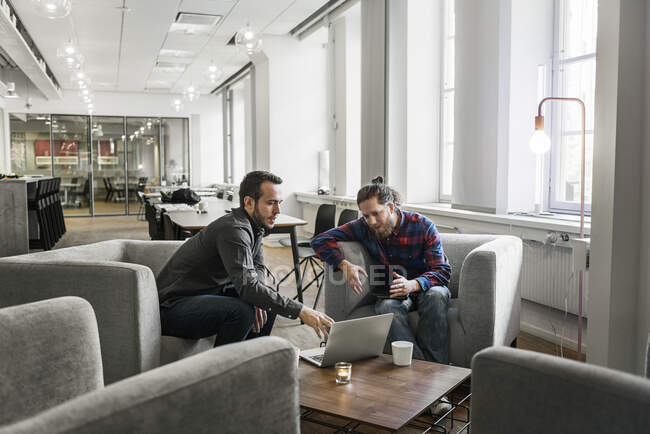 Jovens colegas de trabalho do sexo masculino sentados juntos e usando laptop no escritório — Fotografia de Stock
