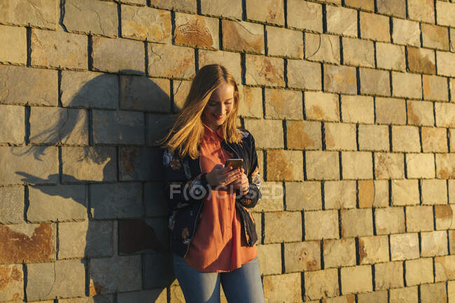 Mulher nova usando telefone inteligente por parede de tijolo — Fotografia de Stock