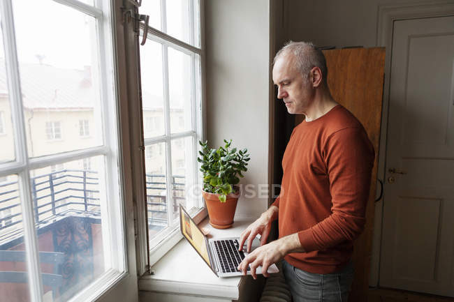 Homme âgé utilisant un ordinateur portable par fenêtre — Photo de stock