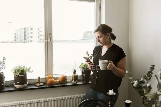 Junge Frau benutzt Smartphone am Fenster — Stockfoto