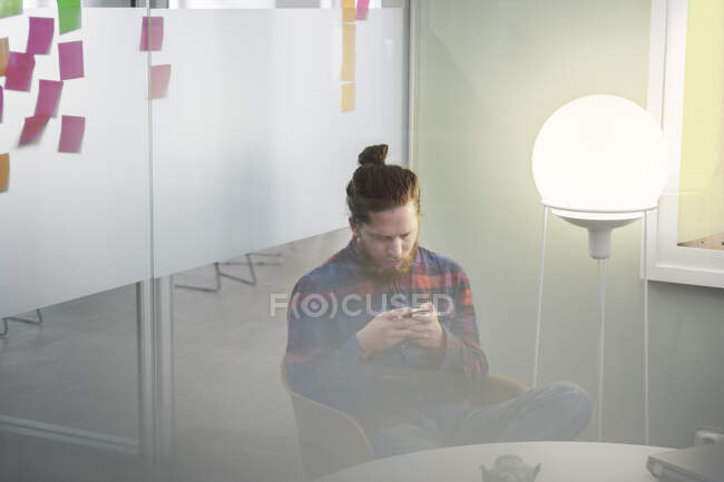 Молодой сосредоточенный человек, сидящий в офисе и пользующийся смартфоном по lamp — стоковое фото