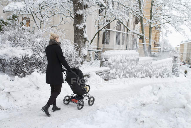 Mujer joven empujando cochecito en la calle de la ciudad nevada - foto de stock