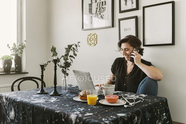 Jeune femme utilisant un ordinateur portable et un téléphone intelligent pendant le petit déjeuner — Photo de stock