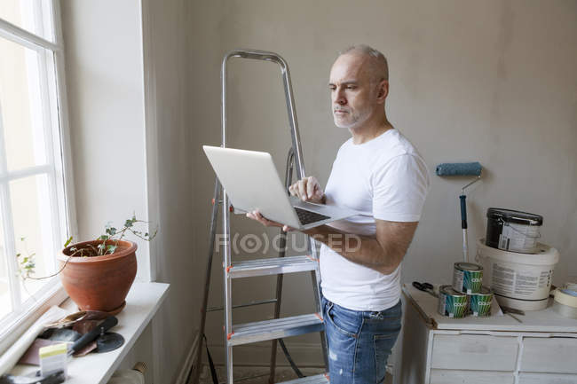 Hombre adulto medio mirando el ordenador portátil durante la renovación del apartamento - foto de stock