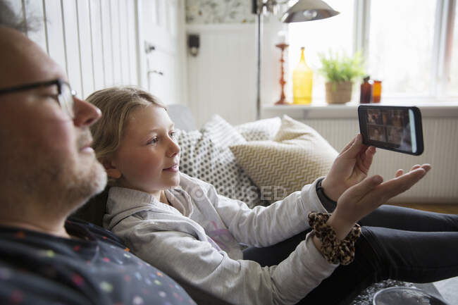 Padre e hija usando un teléfono inteligente mientras están sentados en el sofá - foto de stock