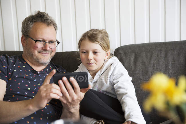 Батько і дочка використовують смартфон, сидячи на дивані — стокове фото