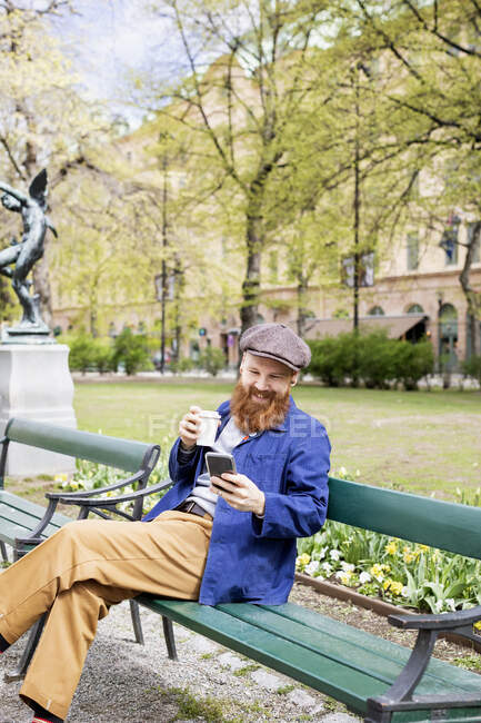 Усміхнений чоловік користується смарт-телефоном на лавці парку. — стокове фото