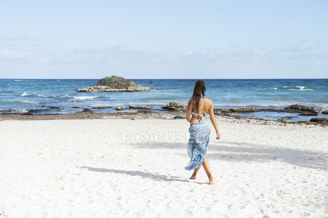 Jeune femme en bikini et sarong marchant sur la plage — Photo de stock