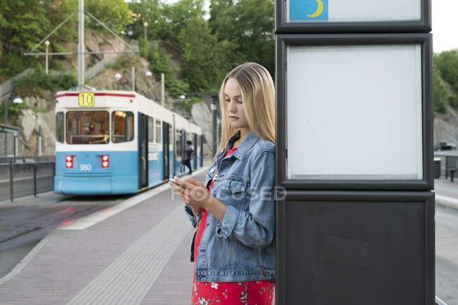 Young woman with smart phone at tram station — Stockfoto
