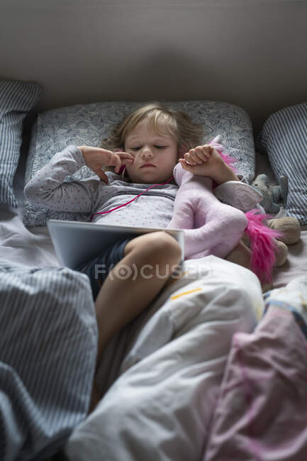 Girl lying in bed with wireless tablet and stuffed toy — Stockfoto