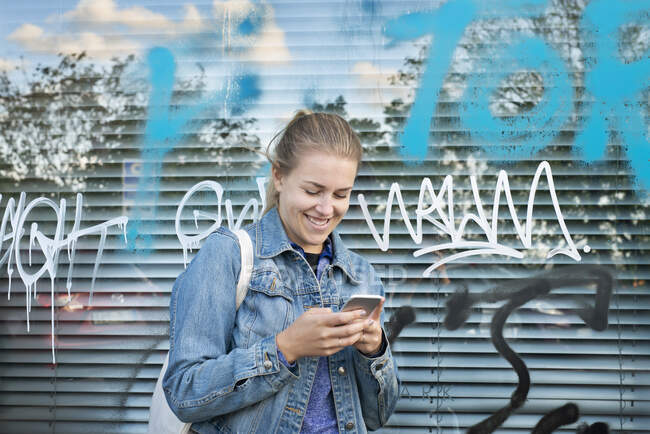 Jeune femme souriante tenant un téléphone portable par fenêtre avec des graffitis — Photo de stock