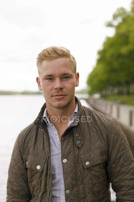 Jeune homme aux cheveux blonds portant une veste au bord du lac — Photo de stock