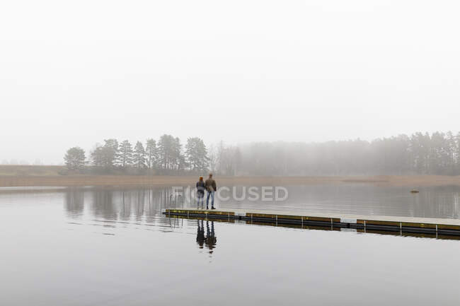 Young man and woman standing on pier at lake - foto de stock