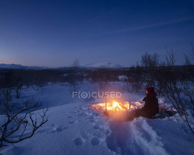 Young woman sitting by campfire in snow — Stock Photo