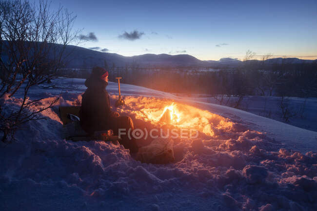 Young woman sitting by campfire in snow — Foto stock