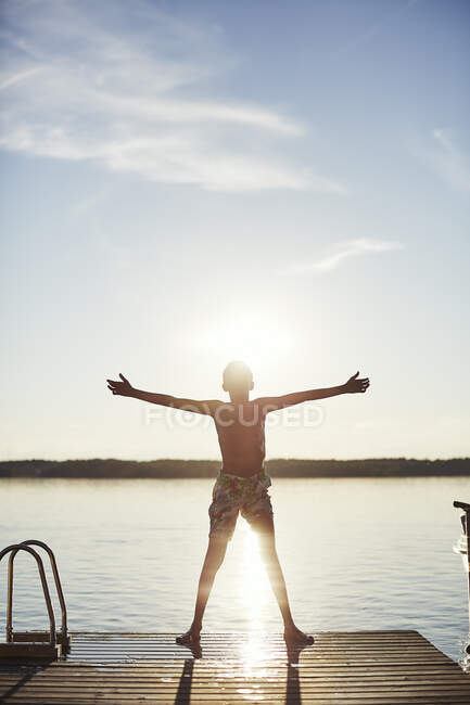 Boy in swimming trunks standing on jetty by lake at sunset — Photo de stock