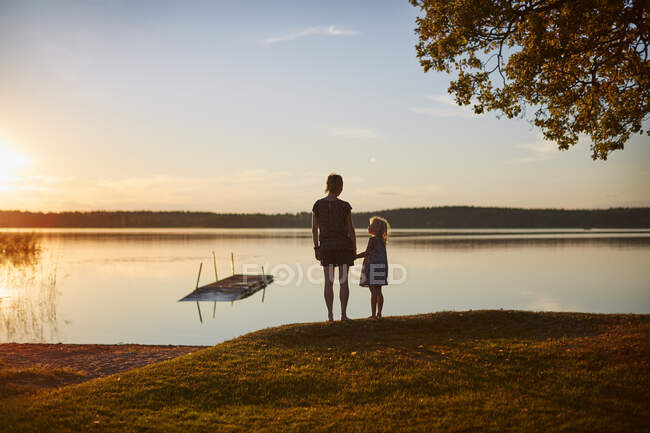 Mujer y su hija de pie junto al lago al atardecer - foto de stock