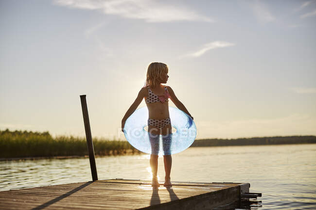 Menina em maiô com brinquedo inflável pelo lago ao pôr do sol — Fotografia de Stock