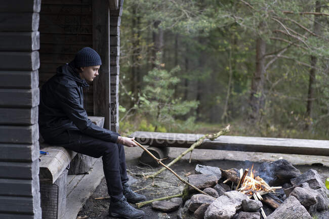 Teenage boy holding stick in campfire by log cabin — Stock Photo