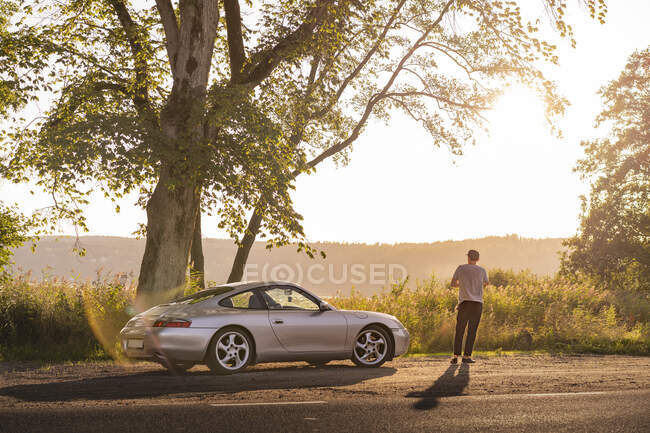 Чоловік стоїть біля спортивної машини на сільській дорозі. — стокове фото