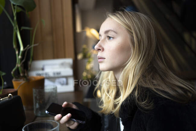 Teenage girl with smart phone looking out window — Stockfoto
