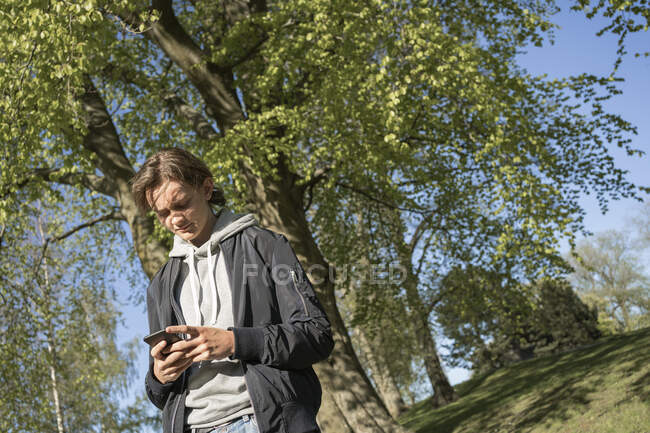 Jovem usando telefone inteligente por árvore — Fotografia de Stock