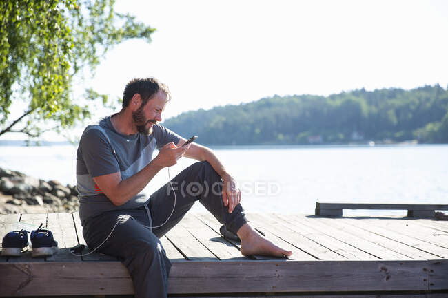 Mann benutzt Smartphone während er auf Steg sitzt — Stockfoto