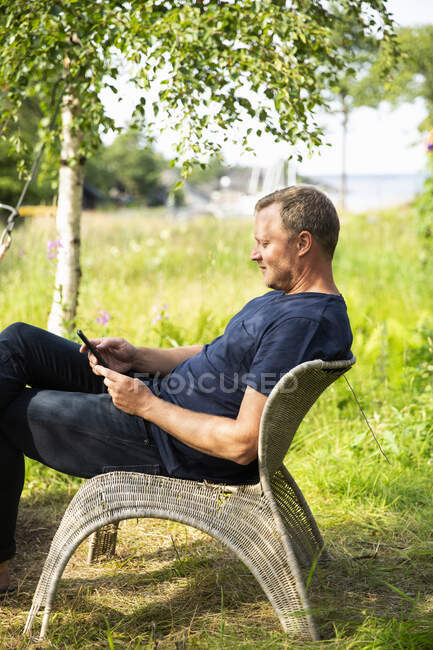 Homem usando telefone inteligente enquanto sentado na cadeira de vime — Fotografia de Stock