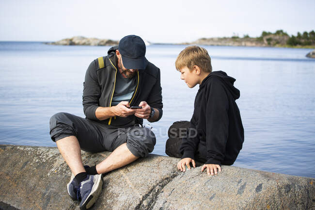Father and son sitting on rock by sea — Stockfoto