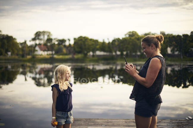 Женщина фотографирует дочь со смартфоном у озера — стоковое фото